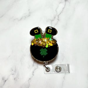 May include: A black pot filled with gold glitter and topped with two green leprechaun boots. The pot has a green shamrock on the front and is attached to a clear plastic badge holder.