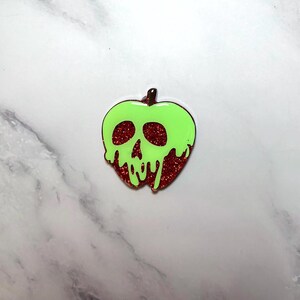 May include: A green apple-shaped pin with a skull design. The apple has a bright green outline and a red glitter interior. The skull's eye sockets and mouth are visible. The pin is a unique accessory.