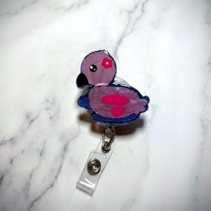 May include: A flamingo-shaped badge reel with a pink body, a black beak, and a small orange flower. The flamingo is outlined in blue glitter and has a clear plastic retractable cord and a metal clip.