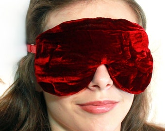 May include: A close-up of a woman wearing a ruby red velvet sleep mask. The mask covers her eyes and is secured with an elastic band.