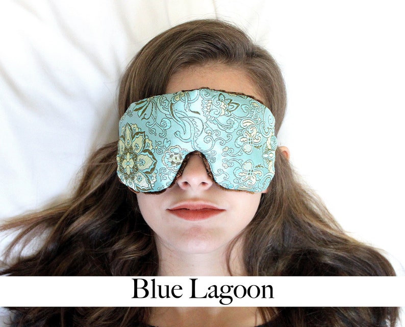 May include: A woman wearing a blue and gold floral eye mask. The eye mask is made of satin and has a comfortable fit. The woman is lying down on a white bed.