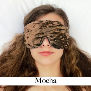 K&ouml;nnte beinhalten: Eine braune Schlafmaske aus Samt mit einem dezenten Schimmer. Die Maske wird von einer Person mit langen braunen Haaren getragen, die auf einer wei&szlig;en Oberfl&auml;che liegt. Das Wort "Mocha" steht unter dem Bild.