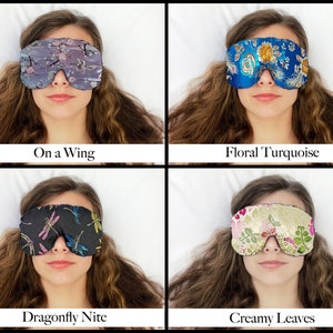 May include: Four sleep masks are shown, each with a unique design. The top left mask is grey with a floral and bird pattern, labelled "On a Wing." The top right mask is blue with a floral pattern, labelled "Floral Turquoise." The bottom left mask is black with dragonfly designs, labelled "Dragonfly Nite." The bottom right mask is cream with a leaf design, labelled "Creamy Leaves."