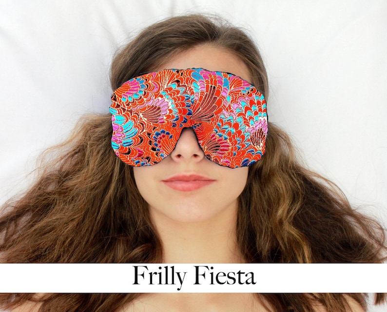 May include: An eye mask with a bright floral design in orange, blue, and pink. The mask is worn over the eyes. The text "Frilly Fiesta" is displayed below the mask.