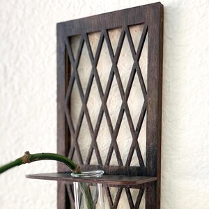 May include: A dark brown wooden wall planter with a diamond lattice design. A clear glass tube holds a green plant with large leaves. The planter has a small shelf to hold the glass tube. The background is a white wall.