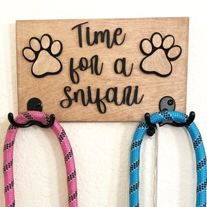 May include: A wooden sign with the text "Time for a sniffari" and two paw prints. Two leashes, one pink and one blue, are hanging on hooks attached to the sign.