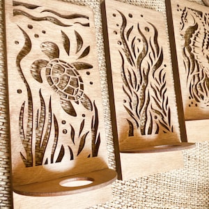 May include: Wooden wall art featuring laser-cut designs of marine life. The art includes a sea turtle, seaweed, and a seahorse. Each piece has a small shelf. The wood has a natural, light brown color.