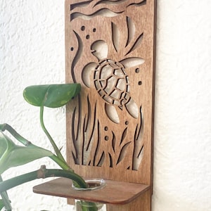 May include: Wooden wall-mounted plant holder with a laser-cut sea turtle design. The brown wooden panel features a turtle, seaweed, and bubbles. A small shelf holds a glass vase with a green plant, creating a decorative and natural accent for any space.