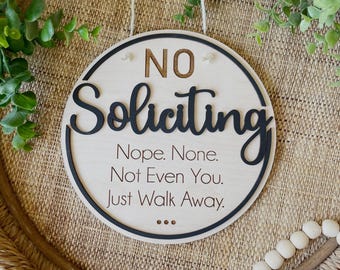 May include: A round wooden sign with the words "NO Soliciting" in black script. The sign also includes the phrases "Nope. None. Not Even You. Just Walk Away." and is designed to hang with a string.