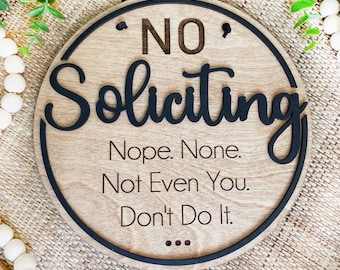 May include: A round wooden sign with the words "NO Soliciting" in black script. Below, the sign reads "Nope. None. Not Even You. Don't Do It." The sign has a black border and a black string for hanging.