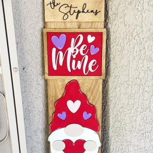 May include: A wooden Valentine's Day gnome door sign with a red hat, white beard, and a red heart in its hands. The sign also features the text "The Stephens", "Be Mine", and "Welcome".
