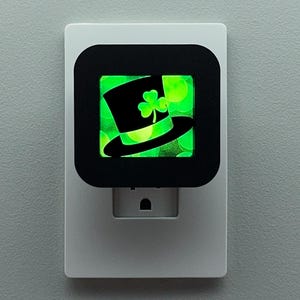 May include: A white night light with a black square face featuring a green glowing image of a black top hat and a shamrock. The night light is plugged into a wall outlet.