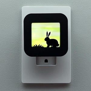 May include: A white night light with a black square frame featuring a silhouette of a rabbit in a field. The background is a gradient of yellow and green. The night light has a plug-in base.