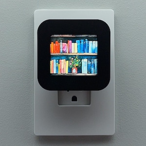 May include: A white night light with a black square display featuring a colorful bookshelf image. The bookshelf is filled with books of various colors, and a small plant is visible on the lower shelf. The night light is plugged into a wall.