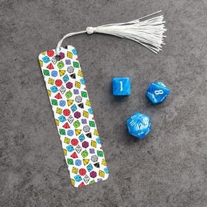 May include: A white bookmark with a colorful pattern of dice. The bookmark has a white tassel. There are four blue dice on a gray surface.