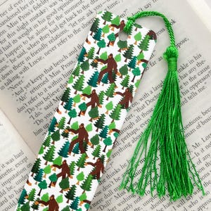May include: A green bookmark with a tassel features a repeating pattern of a brown Bigfoot walking through a forest of green trees on a white background.