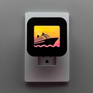 May include: A white night light with a black square face featuring a cruise ship silhouette against a sunset backdrop. The night light is plugged into a gray wall.