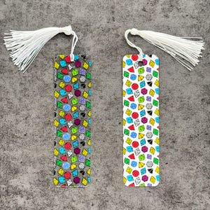 May include: Two white tassel bookmarks with a colorful dice pattern. The bookmarks are made of clear plastic and have a white tassel on each end.