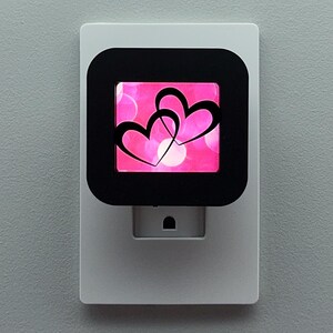 May include: A white night light with a black square frame. The frame displays a pink background with white circles and two black intertwined hearts. The night light is designed to plug into a wall outlet.