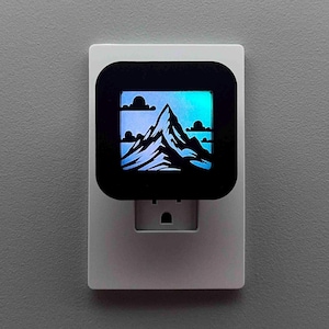 May include: A white night light with a black square face featuring a mountain silhouette design. The mountain is white, with a blue and green gradient background. The night light plugs directly into a wall outlet.