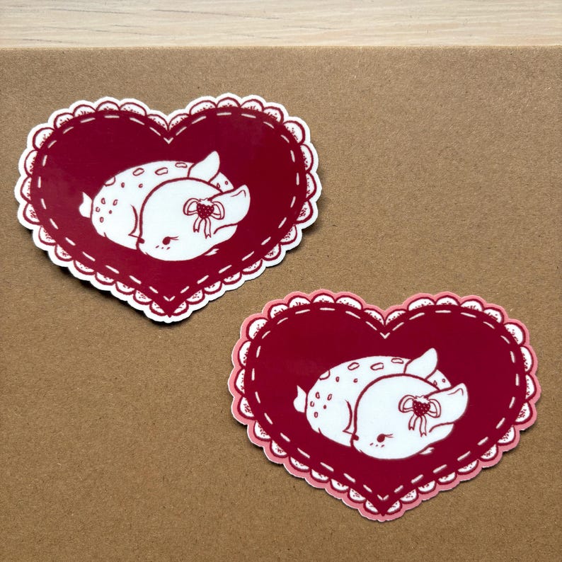 May include: Two red heart-shaped stickers with white outlines and a white deer inside each heart. The deer are lying down with their eyes closed and have a small bow on their head.