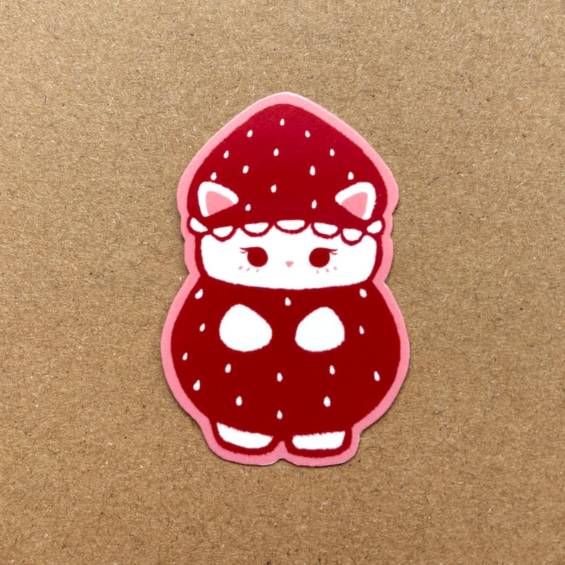 Puede incluir: Un gato blanco con un disfraz de fresa rojo con detalles blancos. El gato tiene un contorno rosa y est&aacute; de pie sobre un fondo marr&oacute;n.