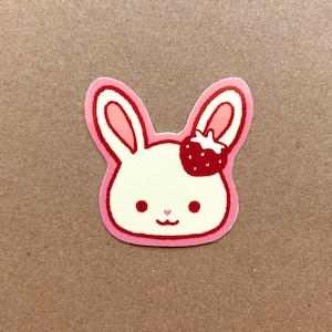 May include: A pink and white sticker of a cartoon bunny with a strawberry on its head. The bunny has a pink nose and pink ears.
