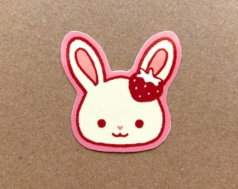 May include: A pink and white sticker of a cartoon bunny with a strawberry on its head. The bunny has a pink nose and pink ears.