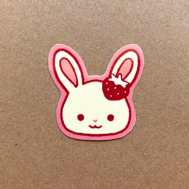 May include: A pink and white sticker of a cartoon bunny with a strawberry on its head. The bunny has a pink nose and pink ears.