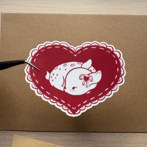 May include: A red heart-shaped sticker with a white outline and a white illustration of a sleeping fawn with a strawberry on its head.