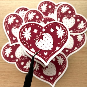 May include: A stack of red heart-shaped stickers featuring a white strawberry and floral design. The stickers are ideal for adding a touch of whimsy to journals, laptops, water bottles, and more.