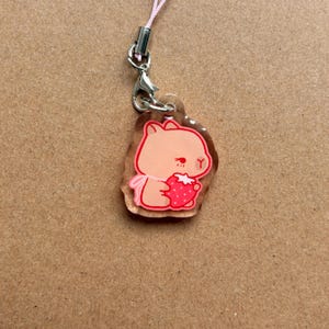 Könnte beinhalten: Ein klarer Acryl-Anhänger mit einem Cartoon-Wombat, der eine Erdbeere hält. Das Wombat ist hellbraun mit einer rosa Schleife, und die Erdbeere ist rot mit weißen Samen. Der Anhänger ist an einer rosa Schnur mit einem silbernen Verschluss befestigt.