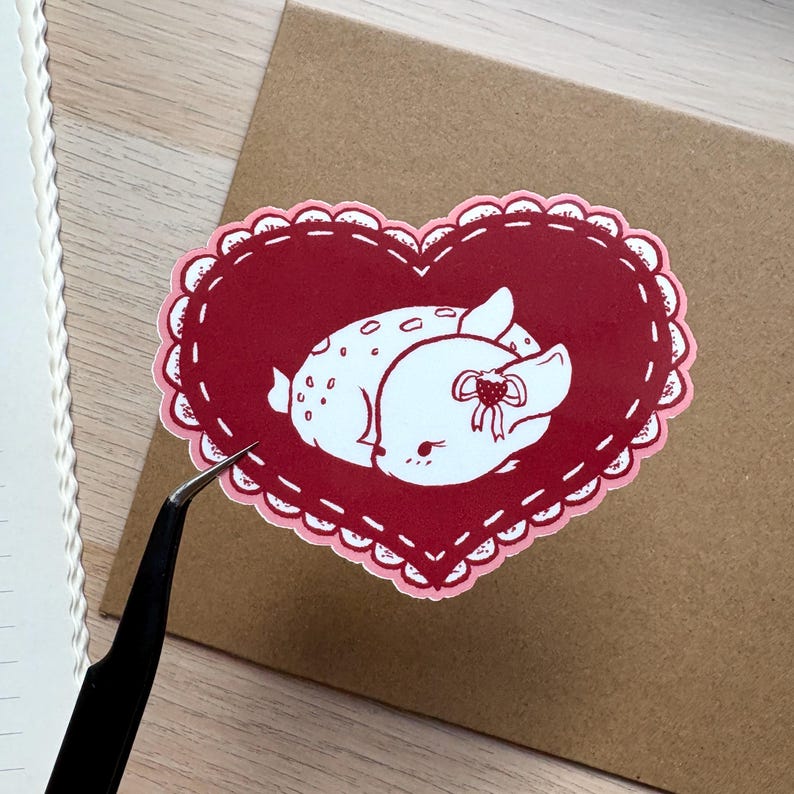 May include: A red heart-shaped sticker with a white outline and a white deer with a red bow inside. The sticker has a scalloped edge.