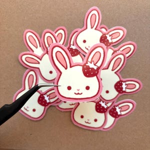 May include: A stack of pink and white bunny stickers with red strawberries on their heads. The stickers are in a cute cartoon style.