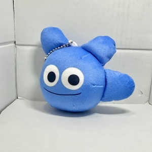 Puede incluir: Un personaje de peluche azul con ojos blancos grandes y pupilas negras. El cuerpo redondo tiene dos pequeños apéndices en forma de ala a cada lado. Una cadena plateada está unida a la parte superior del peluche.
