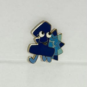 Può includere: Spilla in smalto blu a forma di numero uno con un volto sorridente e due occhi mobili. La spilla ha un bordo dorato e un disegno a stella blu sul lato destro. La spilla è su sfondo bianco.