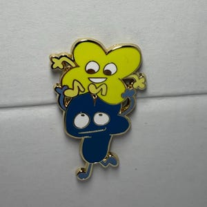 Può includere: Una spilla in smalto color oro con due personaggi dei cartoni animati. Il personaggio in alto è giallo e a forma di lettera "X" con una faccia sorridente. Il personaggio in basso è blu e a forma di numero "4" con un'espressione sorpresa.