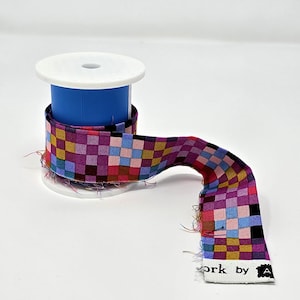 May include: A spool of colorful checkered ribbon with a white and blue spool. The ribbon features a pattern of squares in shades of purple, pink, blue, yellow, and black. The text "work by A" is visible on the end of the ribbon.
