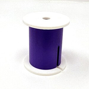 May include: A purple wooden spool with a white top and base. The spool has a cylindrical body with a vertical slot cut into it. The top has a small hole in the center.