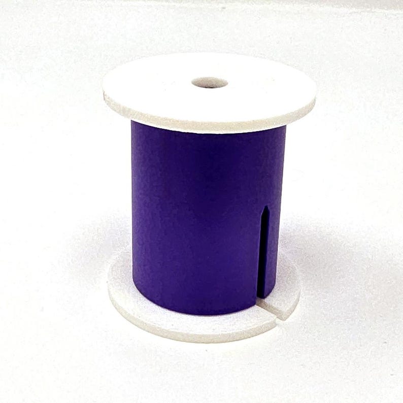 May include: A purple wooden spool with a white top and base. The spool has a cylindrical body with a vertical slot cut into it. The top has a small hole in the center.