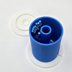 May include: A blue cylindrical container with a white base and lid. The container holds several small blue and black objects. The lid is white and circular. The container is sitting on a white surface.
