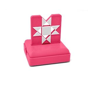 May include: A bright pink square base supports a vertical pink panel featuring a white geometric star pattern. The star design consists of a central square surrounded by eight white triangles arranged in a star shape against the textured pink surface.