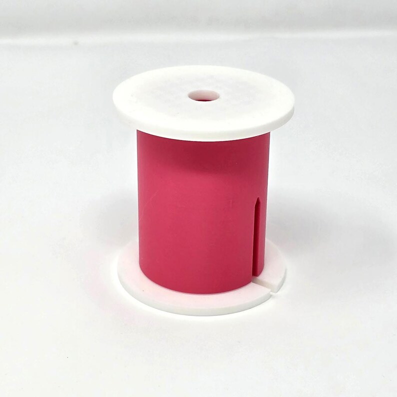 May include: A bright pink spool-shaped object with a white top and base. The cylindrical body has a vertical slot. The top has a small hole in the center. The object is on a white background.