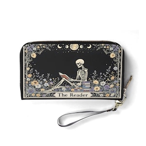 May include: Black wallet with a zipper closure and wrist strap. The front features a skeleton reading a book, surrounded by flowers and moons. The text "The Reader" is below the skeleton.