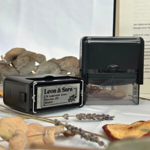May include: A black self-inking stamp with a silver rectangular plate that reads "Leon & Sara 76 Lakeview Drive Denver, CO 80202". The stamp is sitting on a white surface with dried flowers and fruit.