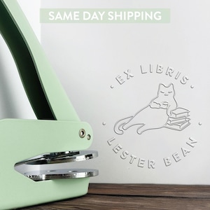 May include: A green self-inking stamp with a silver metal plate. The stamp design features a cat reading a book with the text "Ex Libris Lester Bean".