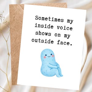 Puede incluir: Una tarjeta de felicitación con el texto "Sometimes my inside voice shows on my outside face." presenta un personaje de dibujos animados azul con una cara sonriente. La tarjeta está sobre un fondo blanco con un sobre marrón.