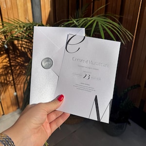 Puede incluir: Una invitación de boda blanca con texto negro y un sello de cera plateado. La invitación incluye los nombres "Cemre & Muratcan" y la fecha "23 Ağustos". La invitación es sostenida por una persona con esmalte de uñas rojo.