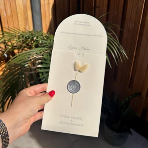 Puede incluir: Una invitación de boda color crema con los nombres "Dylan Mahiou & Sandra Armenak" impresos. La invitación presenta una flor seca y un sello plateado. También es visible el texto "Mr et Mme Armenak" y "Ost la joie de vous faire part du mariage de leurs enfants".
