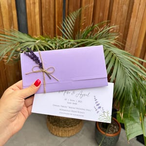 Op de afbeelding: Een lila trouwuitnodiging met bijpassende envelop, vastgebonden met touw en een takje lavendel. De uitnodiging toont de namen "Miso & Aysel" en evenementdetails. De achtergrond bevat houten latten en bladeren.